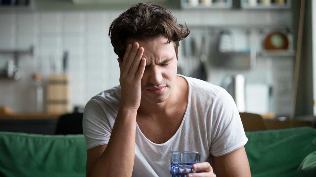 Bilde av mann som sitter i sofaen med et glass vann i den ene hånden og holder seg til hodet med den andre hånden